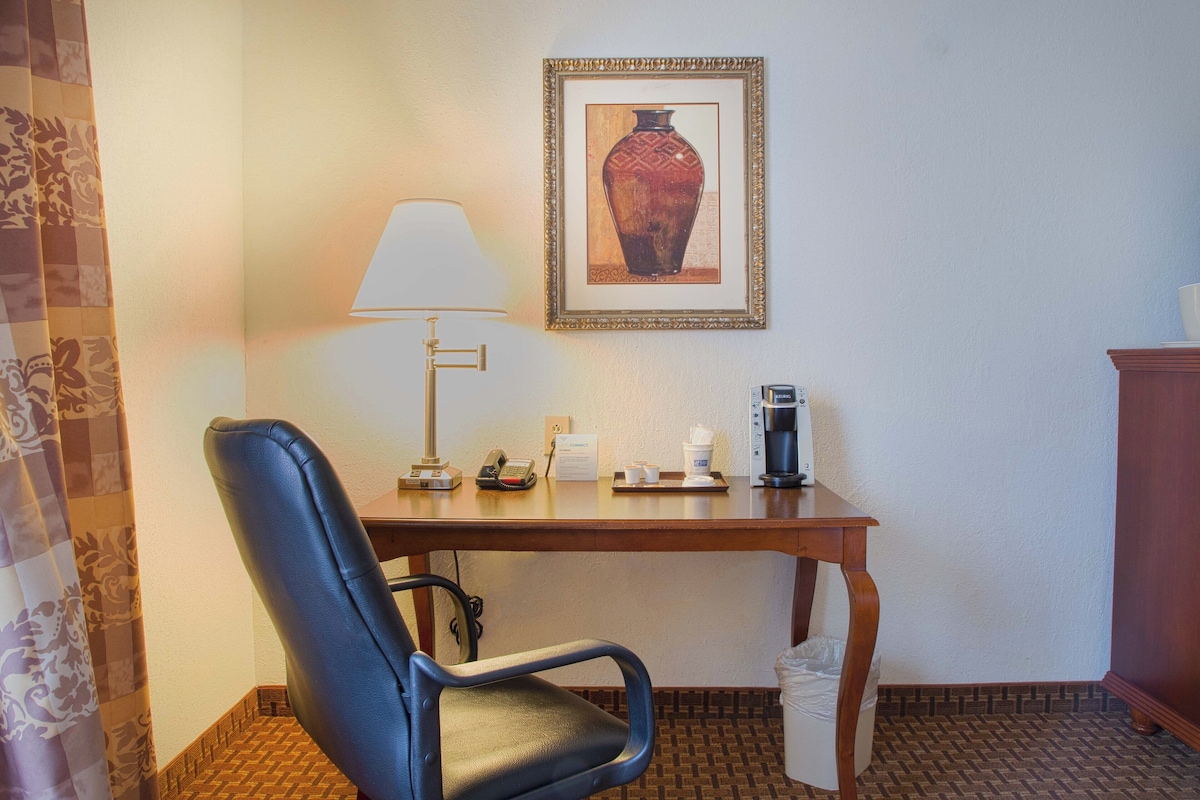 Et hotelværelse med skrivebord, stol, lampe, et indrammet billede, kaffemaskine og skraldespand.