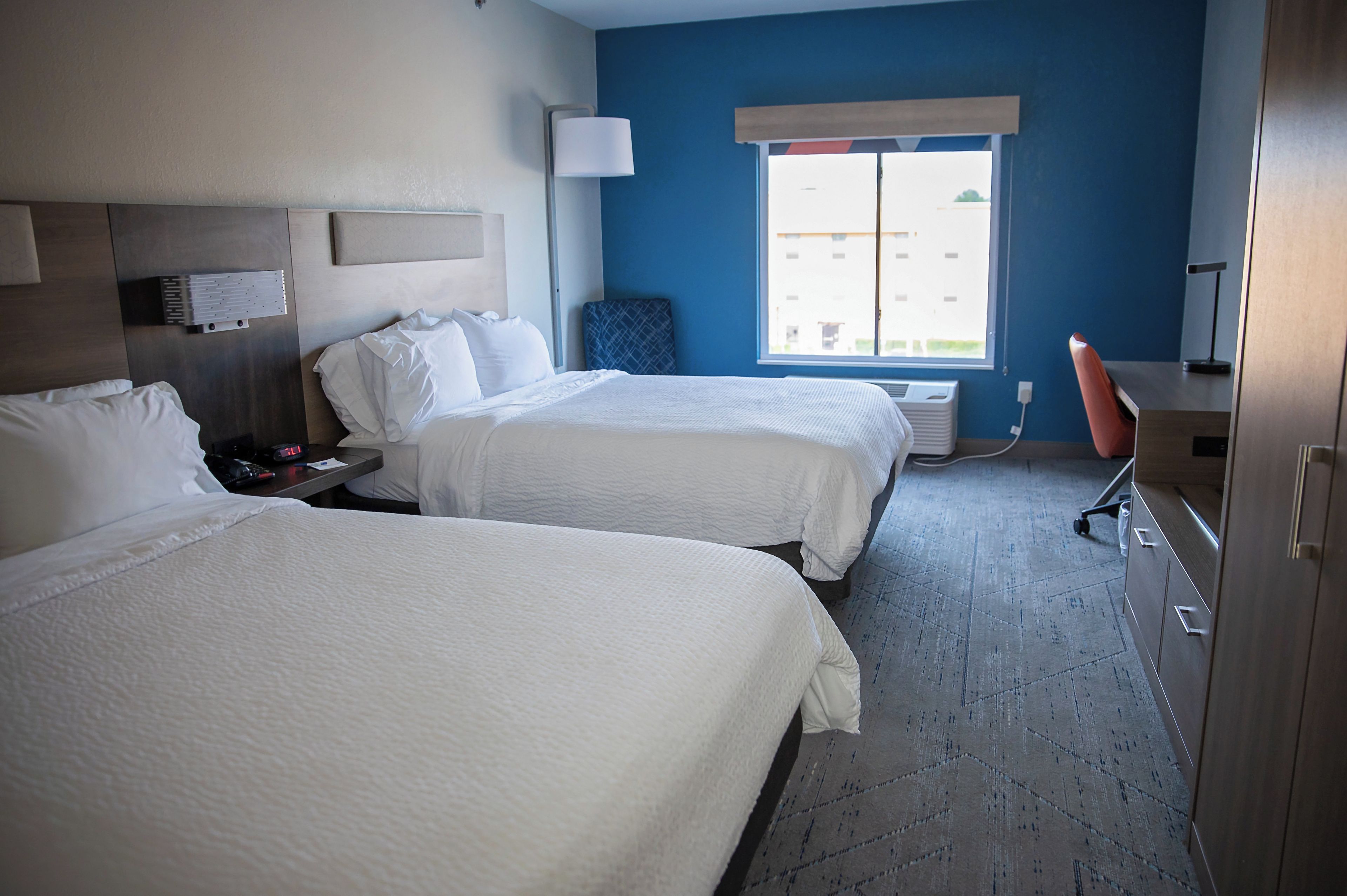 A hotel room with two beds, a desk, a chair, a lamp, and a window with a view.