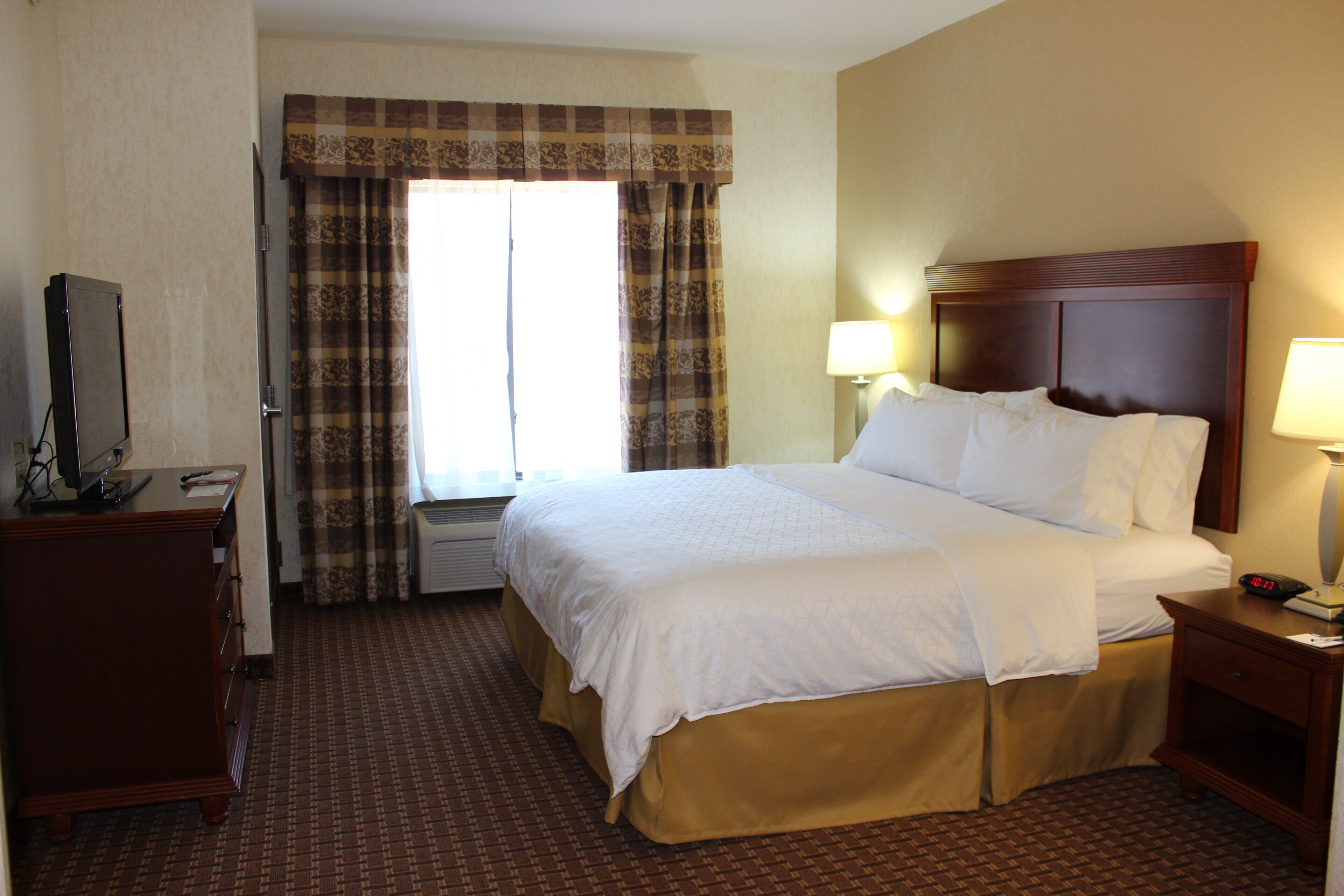 A hotel room with a large bed, a dresser, a television, and a window with curtains.