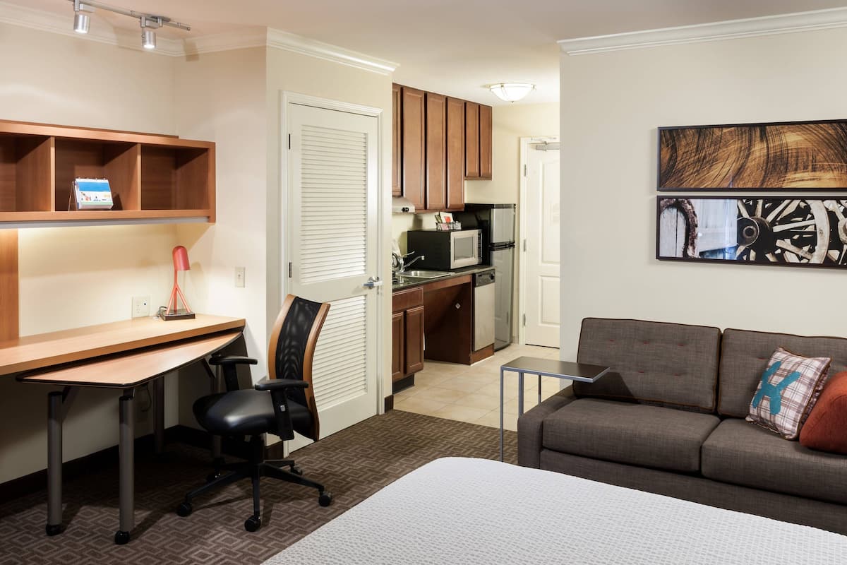 Une chambre d’hôtel compacte comprenant un lit, un bureau avec une chaise, une kitchenette et un canapé gris avec un coussin.
