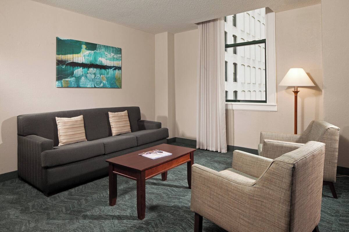 Une chambre d’hôtel avec un canapé gris foncé, une table basse en bois, un fauteuil beige, une lampe sur pied et un tableau au mur.