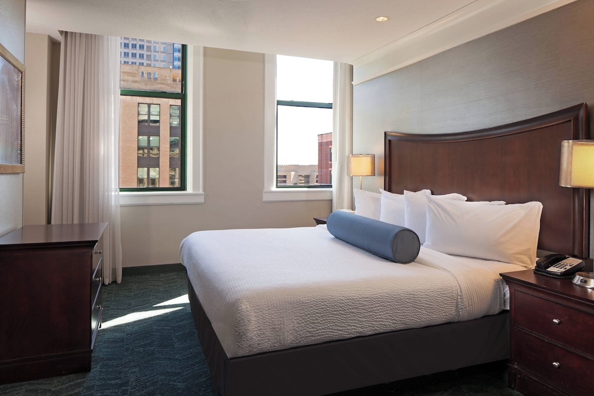 Une chambre d’hôtel avec un grand lit, une table de chevet avec une lampe, une commode et une fenêtre avec des rideaux.