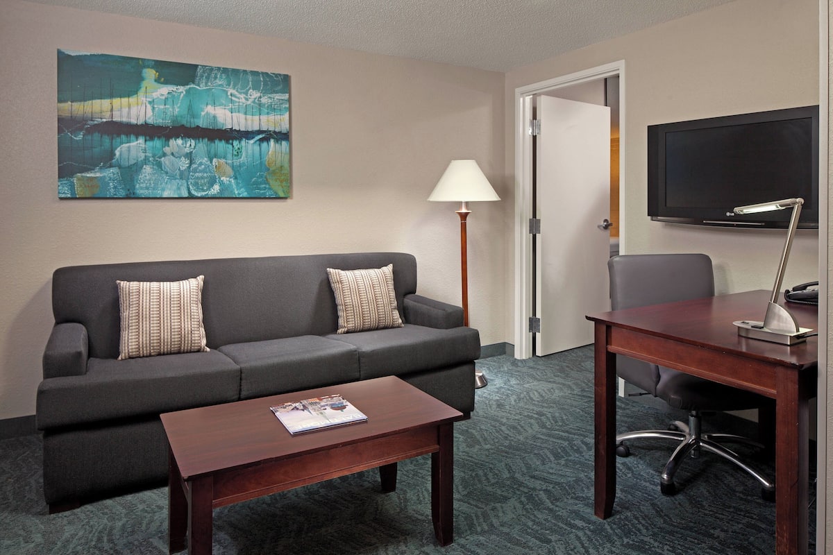 Une chambre d’hôtel avec un canapé gris, une table basse en bois, un bureau avec une chaise, une lampe et un téléviseur à écran plat fixé au mur.