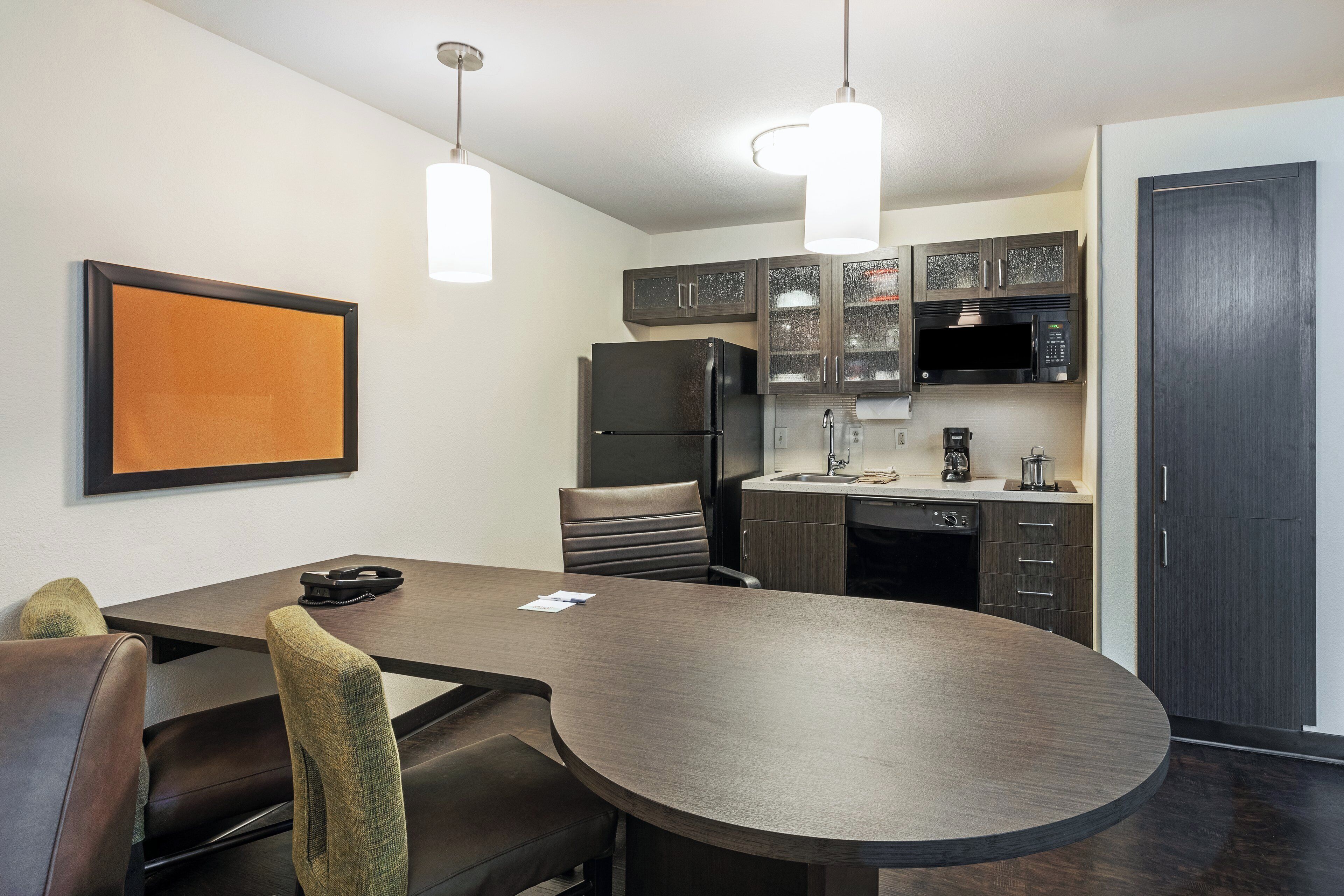 A modern kitchen with dark cabinetry, a black refrigerator, a microwave, and a coffee maker. There is a round table with chairs and a framed picture on the wall.