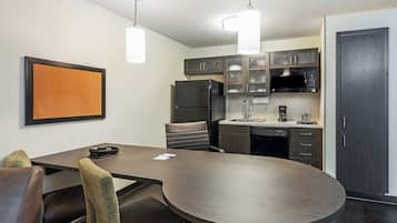 A modern kitchen with dark cabinetry, a black refrigerator, a microwave, and a coffee maker. There is a round table with chairs and a framed picture on the wall.