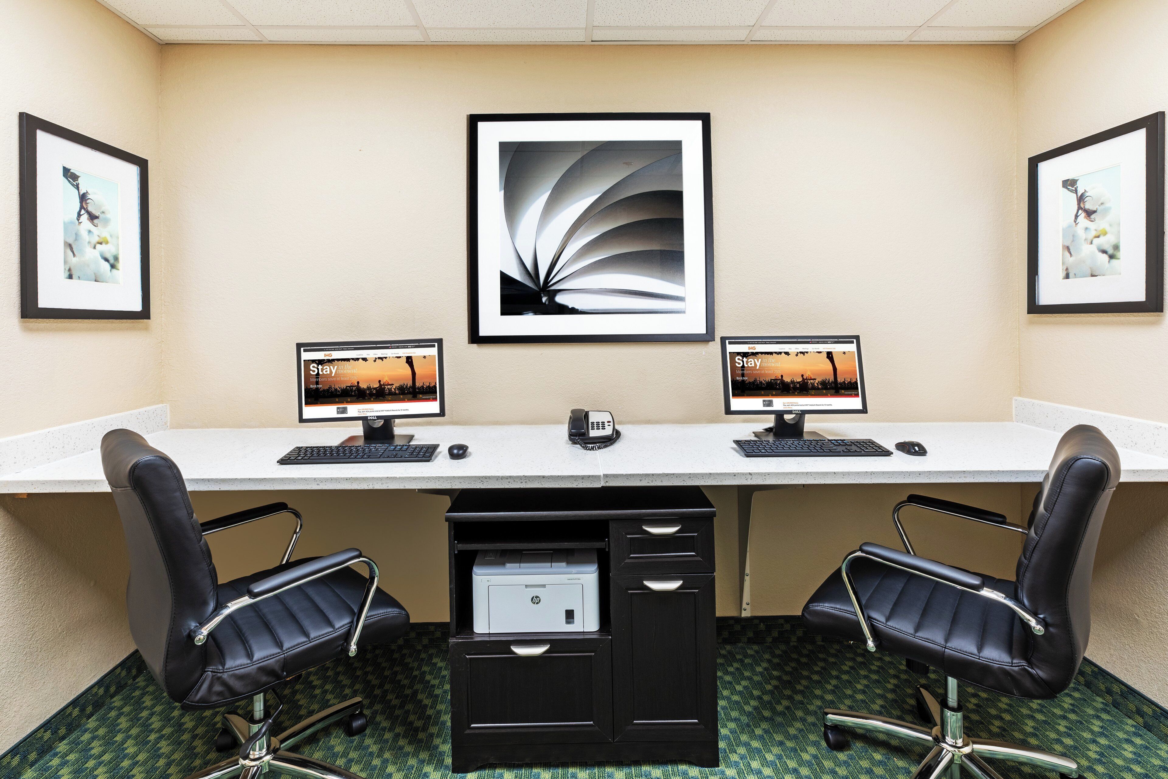 A conference room with two desks, each with a computer, keyboard, and mouse. There are two chairs, a printer, and framed pictures on the wall.