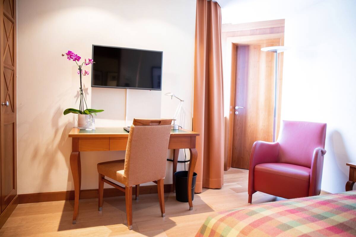 Ein Hotelzimmer mit Holztisch, einem Sessel, einem Fernseher, einem pinken Sessel und einem Bett mit bunter Decke.
