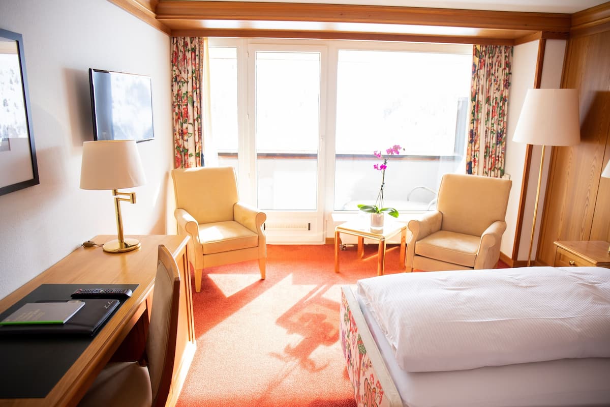Ein Hotelzimmer mit einem Bett, zwei Sesseln, einem Schreibtisch, einer Lampe, einem kleinen Tisch mit einer Pflanze und einem Fenster mit Vorhängen.