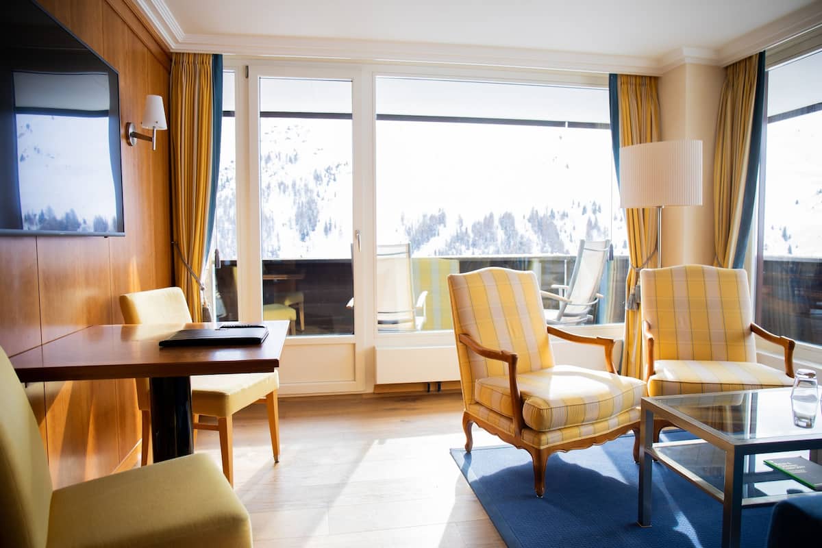 Ein Hotelzimmer mit Holzvertäfelung, einem Esstisch, zwei Sesseln, einer Couch, einem Glastisch und einem großen Fenster mit Blick auf einen verschneiten Berg.