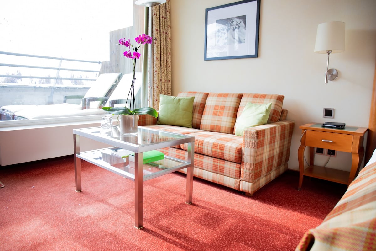 Ein Hotelzimmer mit einem karierten Sofa, einem Glastisch, einem kleinen Nachttisch und einem Fenster mit Vorhängen.