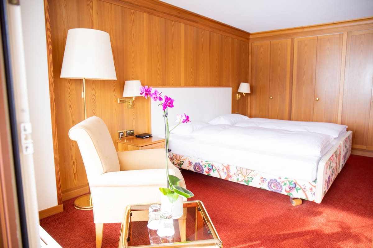 Ein Hotelzimmer mit einer holzverkleideten Wand, einem Bett mit floralem Bettbezug, einem weißen Sessel, einem Nachttisch mit Lampe und einer Vase mit Blumen.