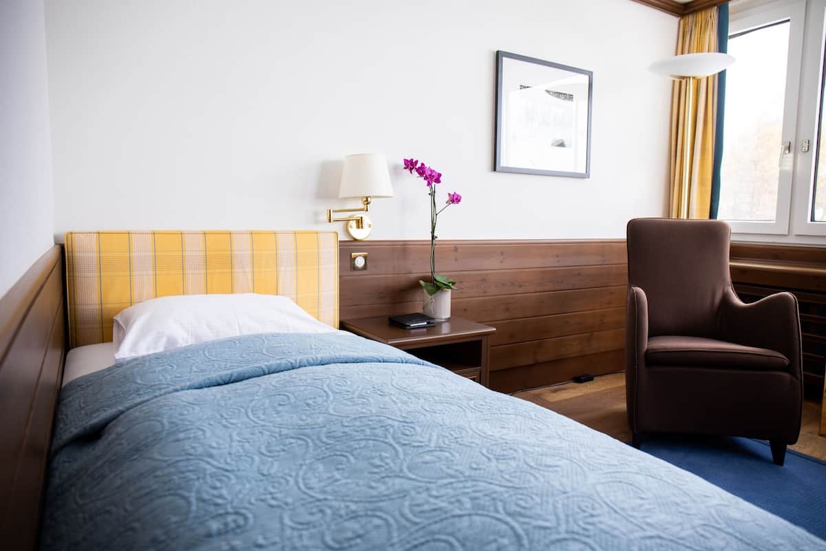 Ein Hotelzimmer mit einem Bett, einem Schreibtisch, einem Stuhl, einem Nachttisch, einer Lampe, einem gerahmten Bild und einem Fenster mit Vorhängen.
