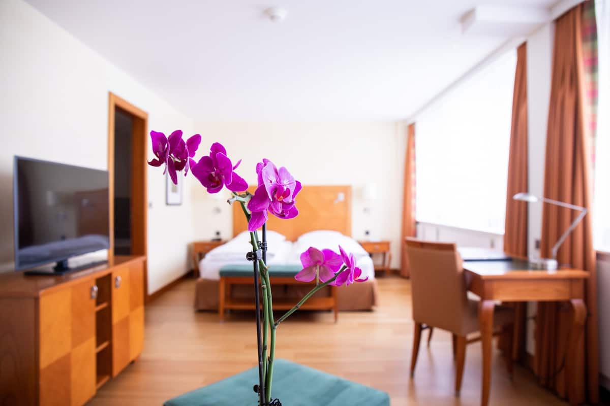 Ein Hotelzimmer mit einem Bett, einem Schreibtisch, einem Fernseher und einer Vase mit violetten Blumen.