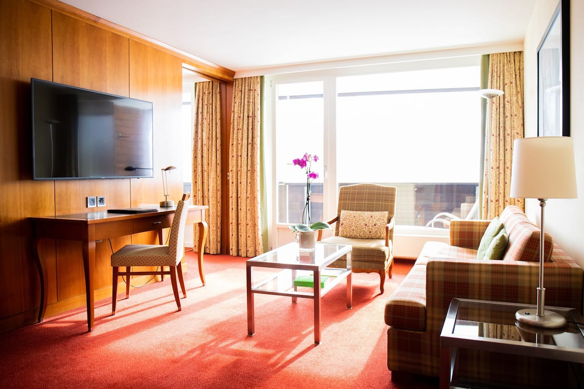 Ein Hotelzimmer mit einem Holztisch, einem Fernseher, einer Couch, einem Sessel und einem großen Fenster mit Vorhängen.