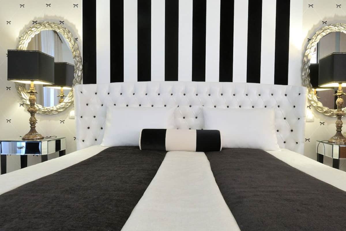 Un dormitorio con un cabecero capitonado, papel tapiz a rayas blancas y negras, y dos mesitas de noche con lámparas.