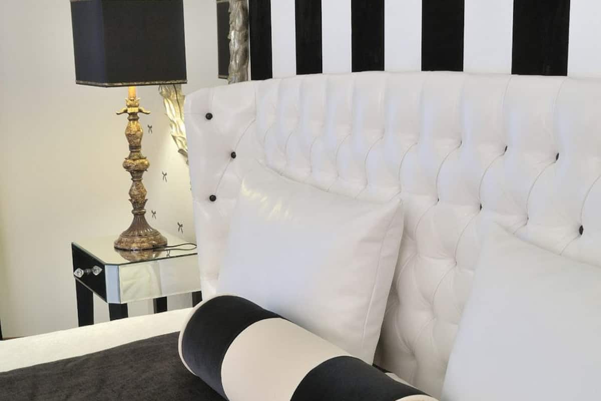 Una cama con cabecera blanca, pared a rayas negras y blancas, y una lámpara sobre una mesita de noche.