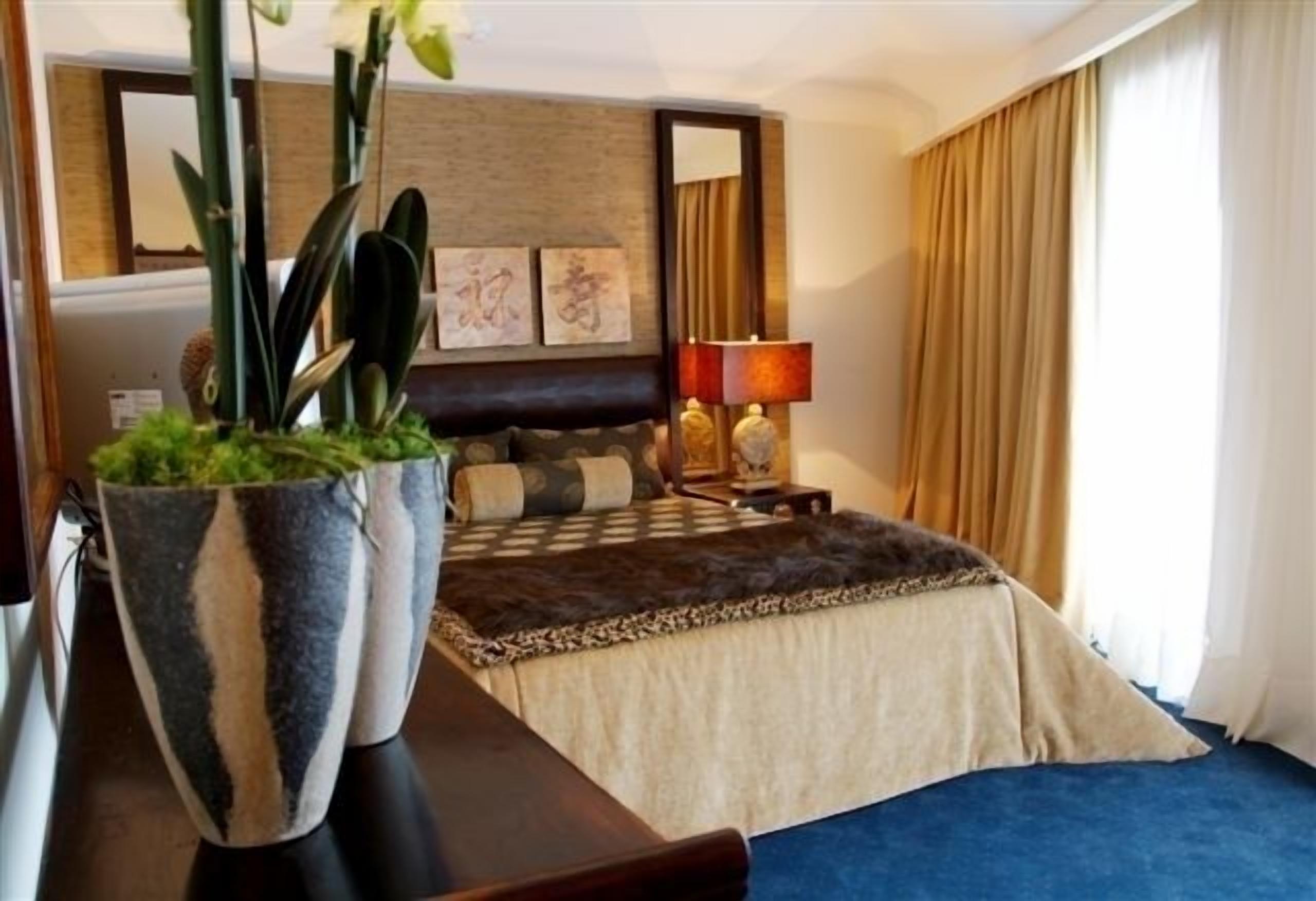 A hotel room with a bed, a large potted plant, a desk, a lamp, and a window with curtains.