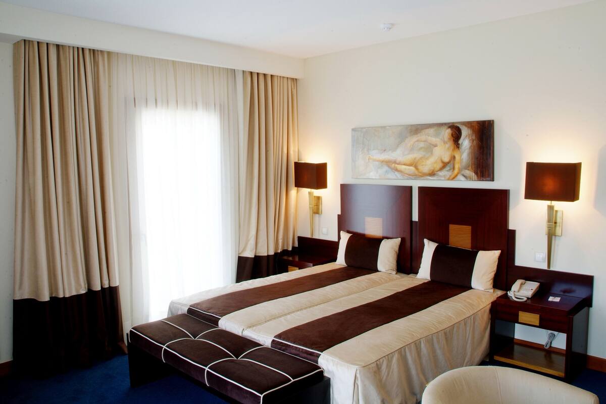 Una habitación de hotel con una cama grande, dos mesitas de noche, un buró, un cuadro en la pared y una ventana con cortinas.