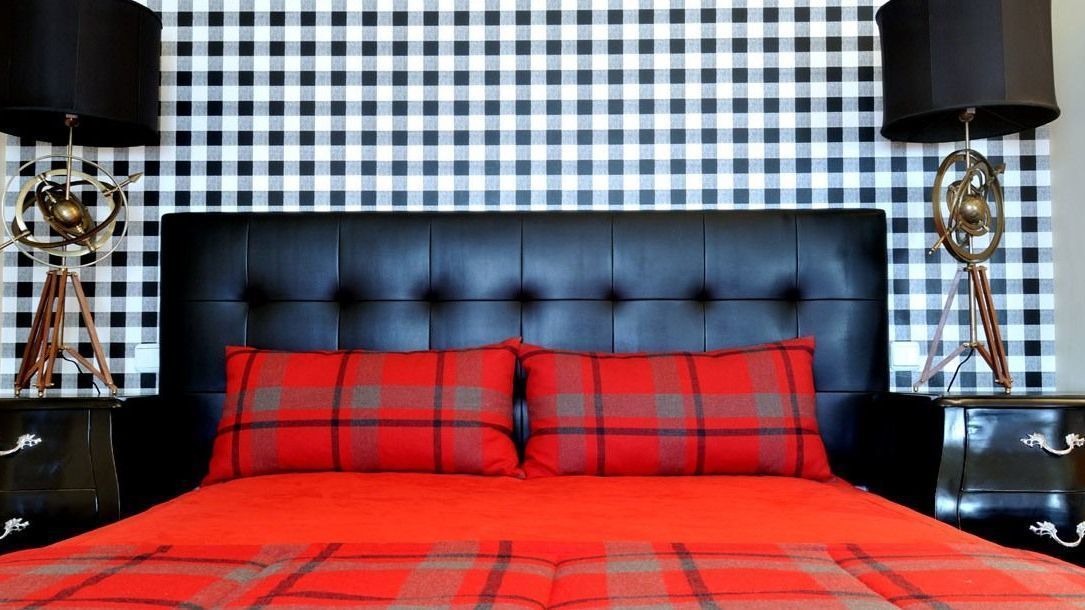 Een bed met een rood-grijs geruite spreideken, een zwart bekleden hoofdeinde en twee zwarte nachtkastjes met lampen.