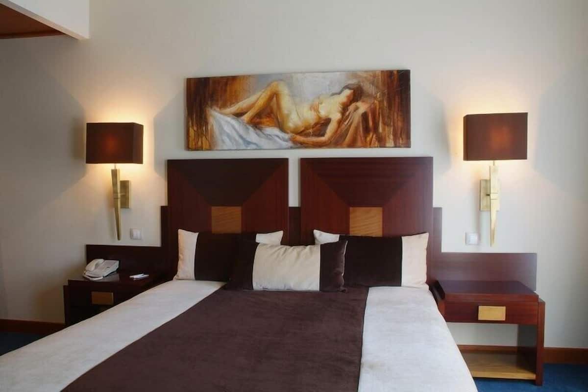 Una habitación de hotel con una cama grande, dos mesitas de noche, un cuadro sobre la cama y lámparas fijadas a la pared.