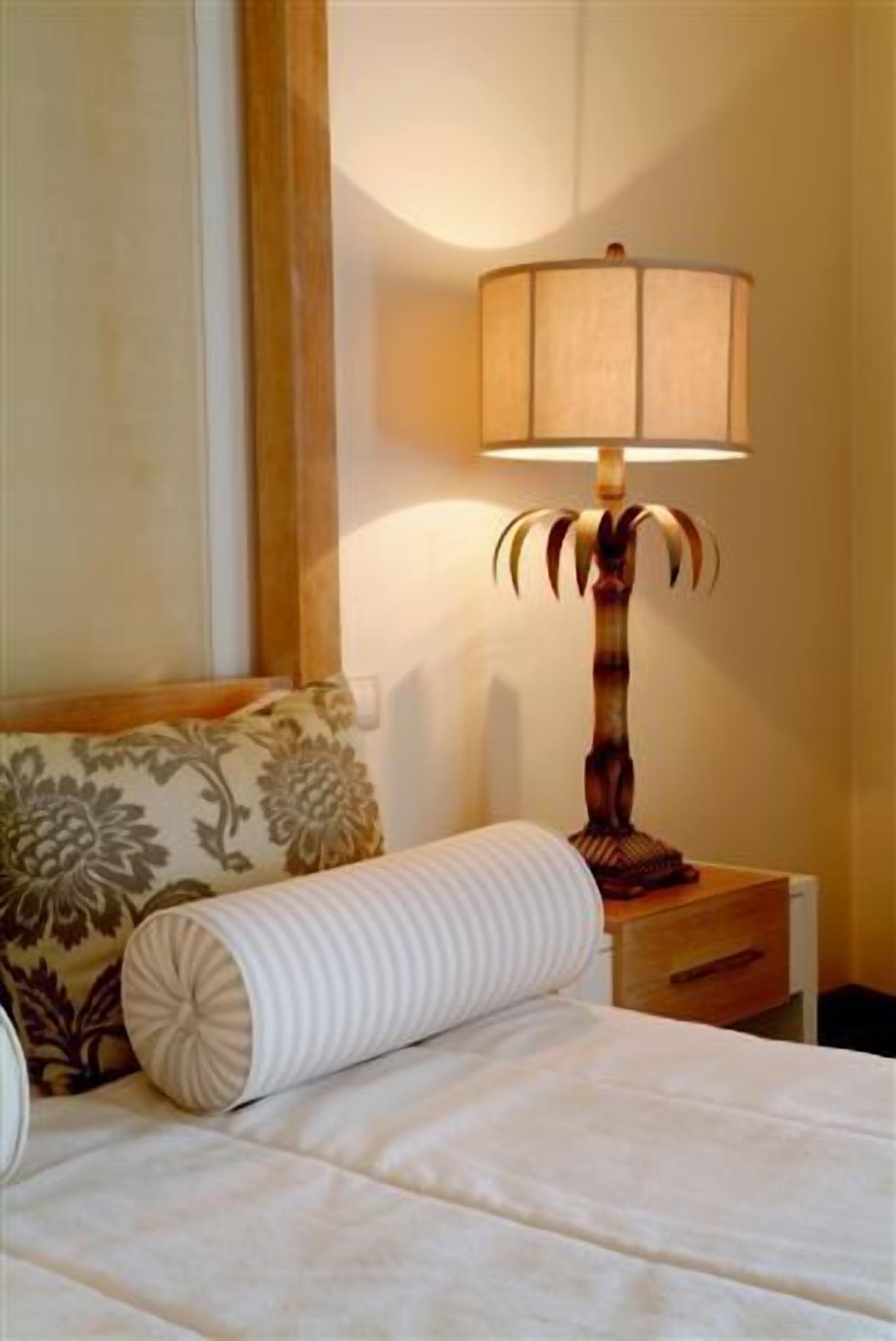 A neatly made bed with a striped pillow and a floral-patterned pillow, a wooden nightstand with a lamp, and a wall with a curtain.