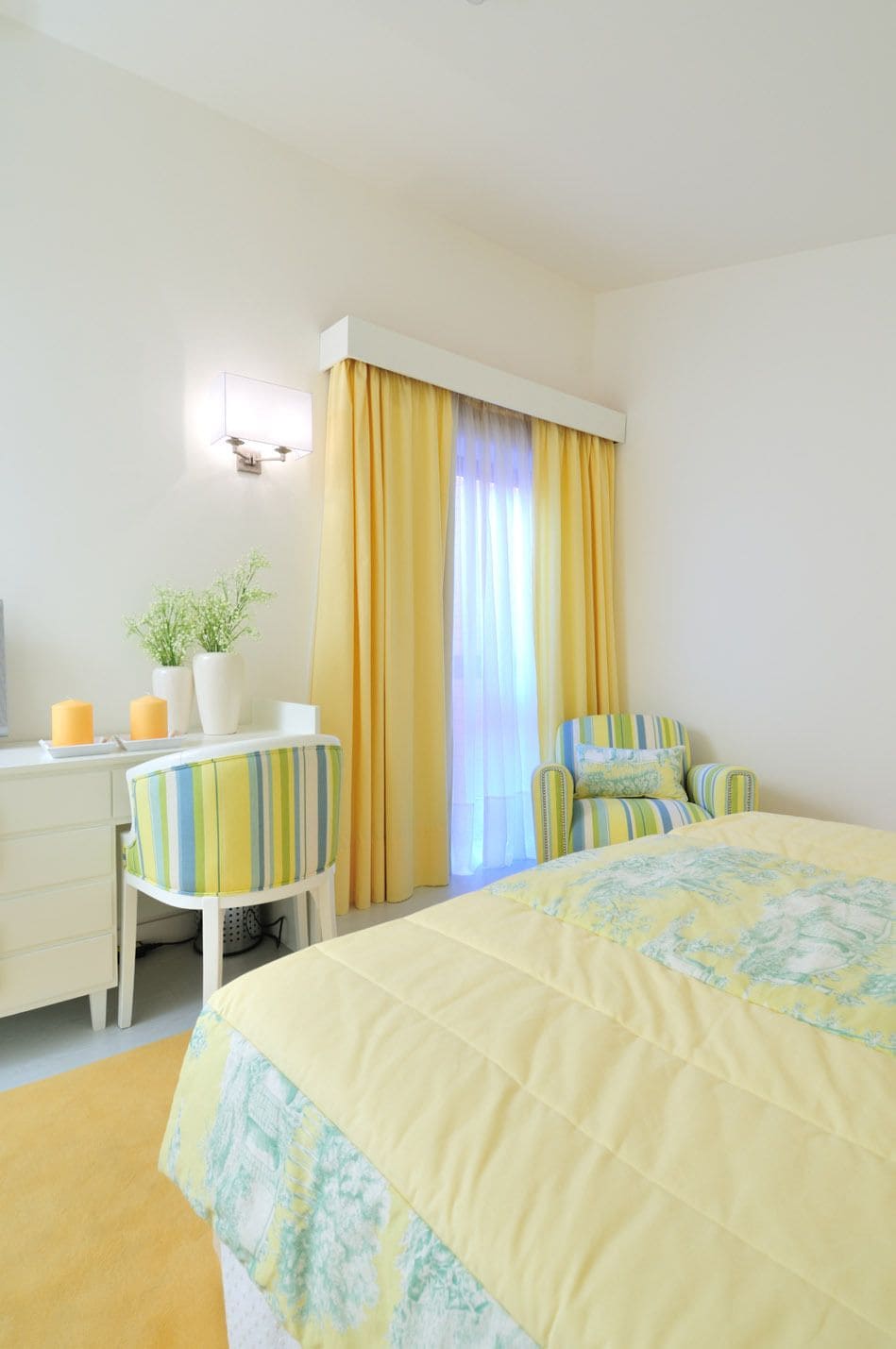 A hotel room with a bed, a desk, a chair, a potted plant, and a window with curtains.