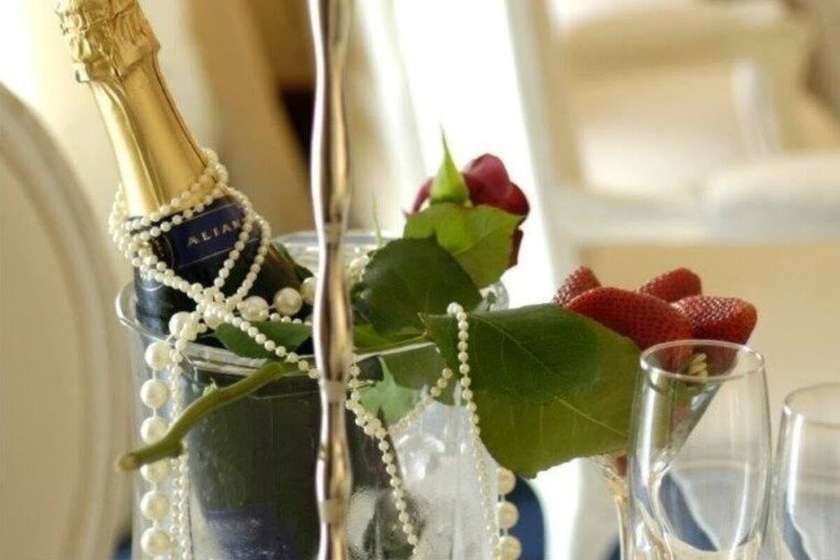 Una botella de champán en una copa con perlas y una rosa, acompañada de fresas y otra copa.