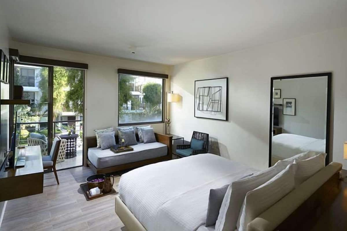 Une chambre d’hôtel moderne, dotée d’un grand lit, d’un canapé, d’un bureau et d’une chaise. Une fenêtre offre une vue sur l’extérieur, et un miroir est accroché au mur.