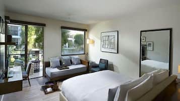A modern hotel room with a large bed, a sofa, a desk, and a chair. There is a window with a view of the outside, and a mirror on the wall.