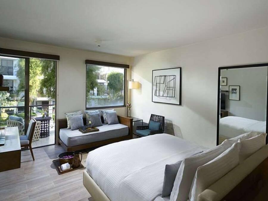 A modern hotel room with a large bed, a sofa, a desk, and a chair. There is a window with a view of the outside, and a mirror on the wall.