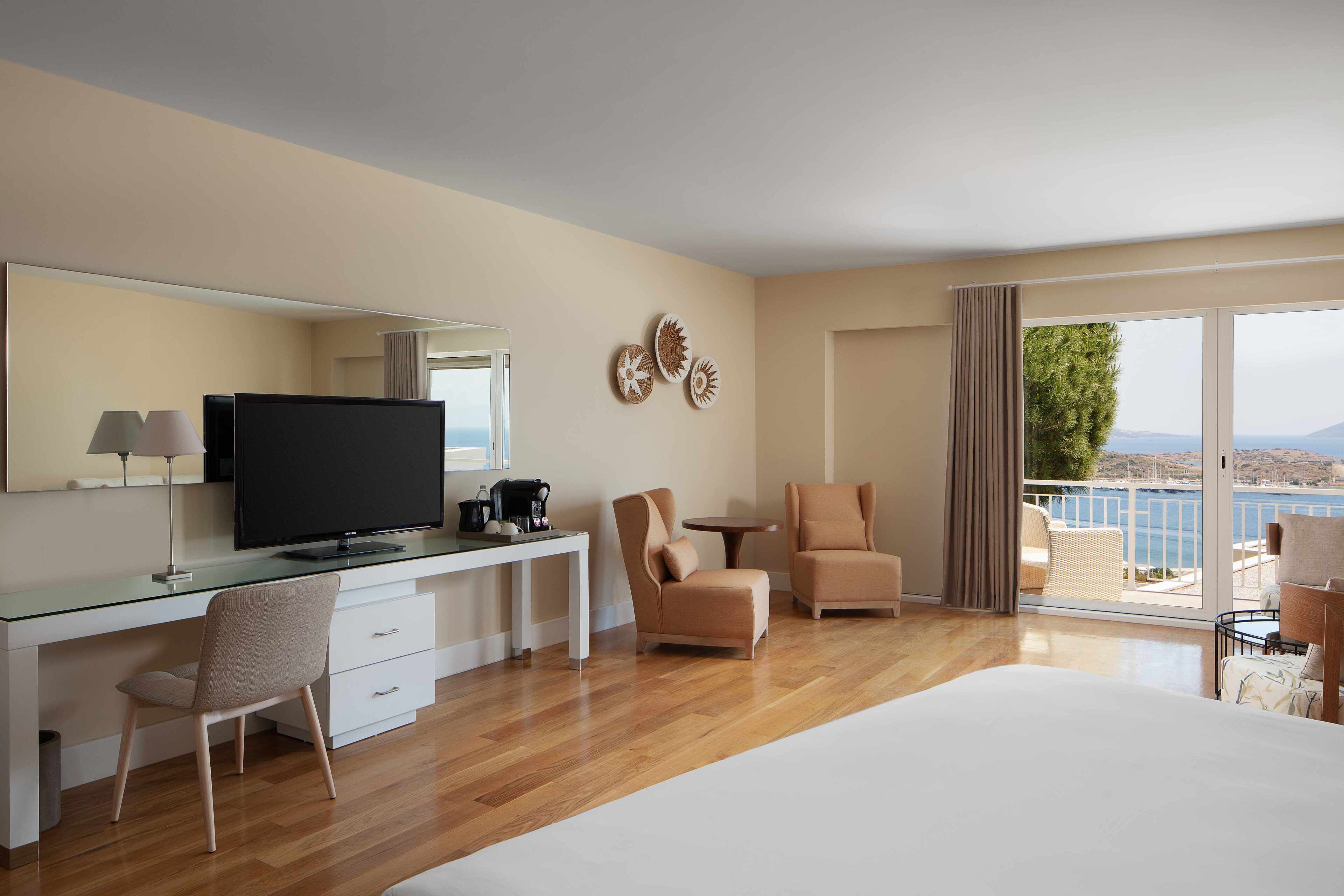 A modern hotel room with a large bed, a desk with a flat-screen TV, two armchairs, a small round table, and a balcony with a view of the sea.