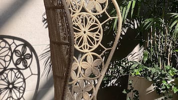 Uma cadeira de vime com desenhos elaborados, posicionada em frente a uma parede com a sombra de uma peça de metal decorativa.
