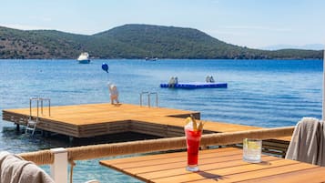 Terraza de madera con una mesa y una bebida, con vistas a un lago tranquilo con barcos y una colina distante.