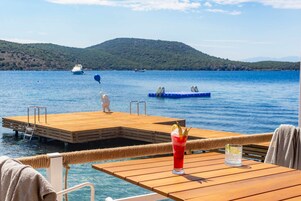 Terraza de madera con una mesa y una bebida, con vistas a un lago tranquilo con barcos y una colina distante.