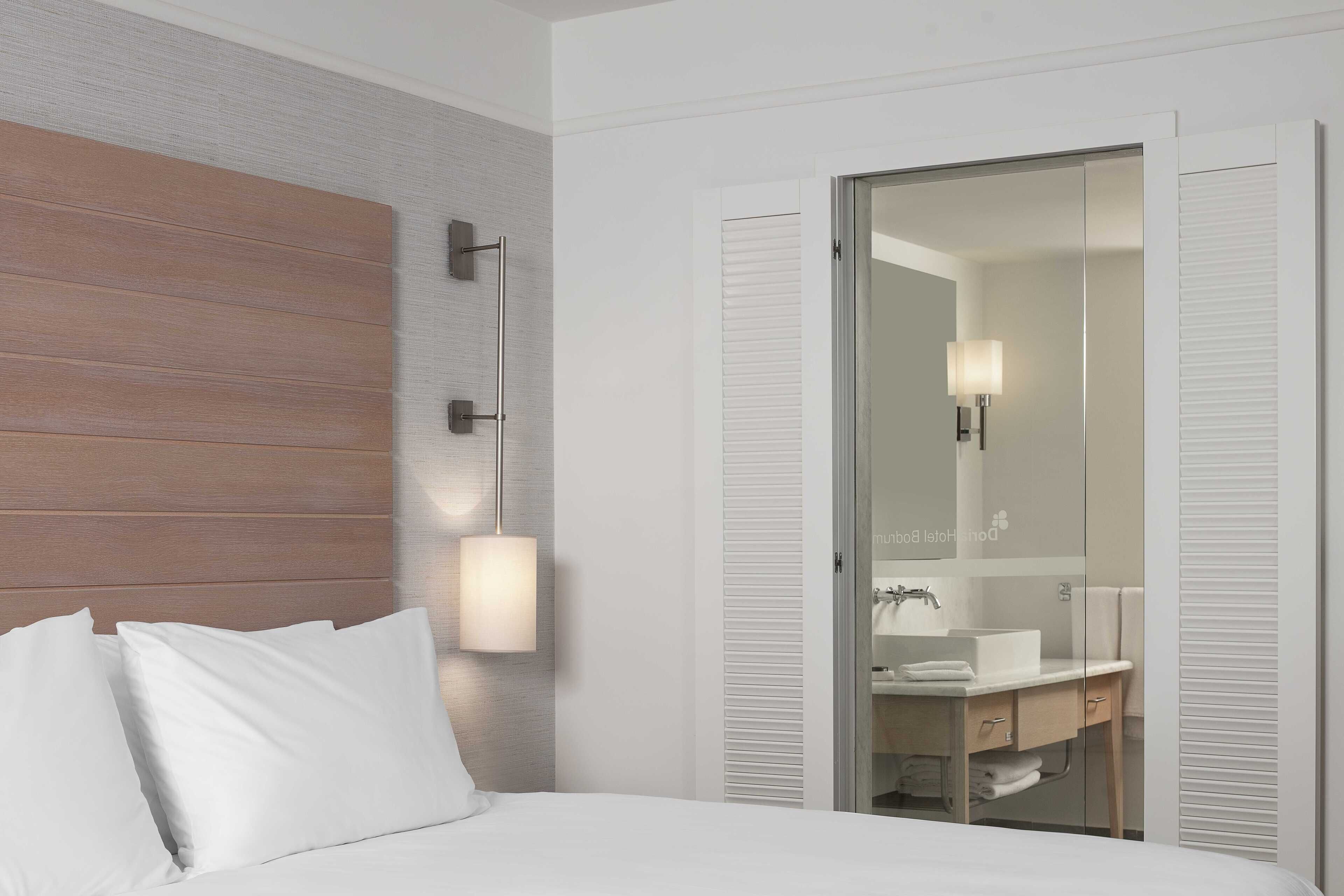 A hotel room with a bed, a wooden headboard, a wall-mounted light, a glass door leading to a bathroom with a sink and mirror.