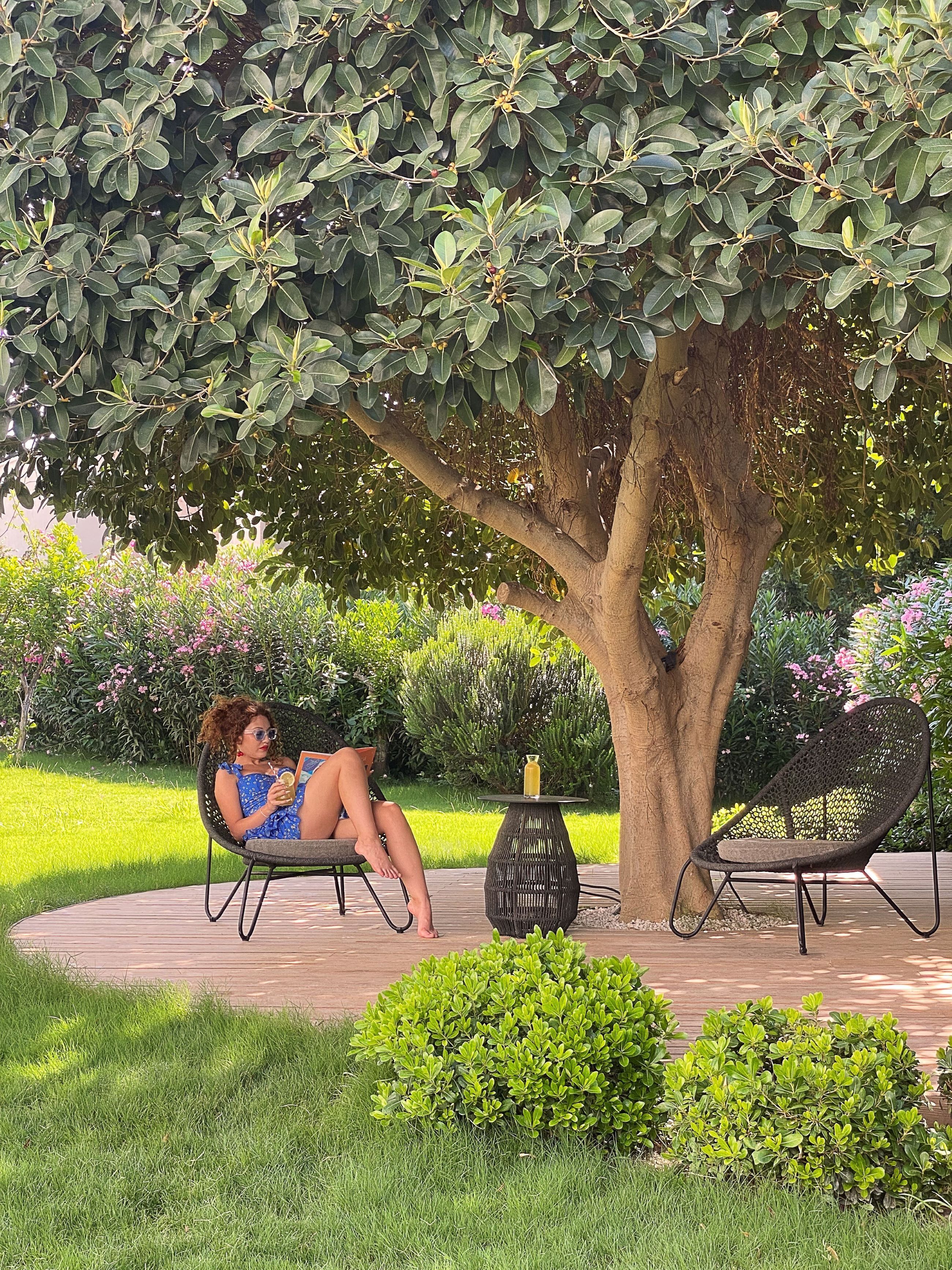 A woman relaxing on a chair under a tree, with a table and another chair nearby.