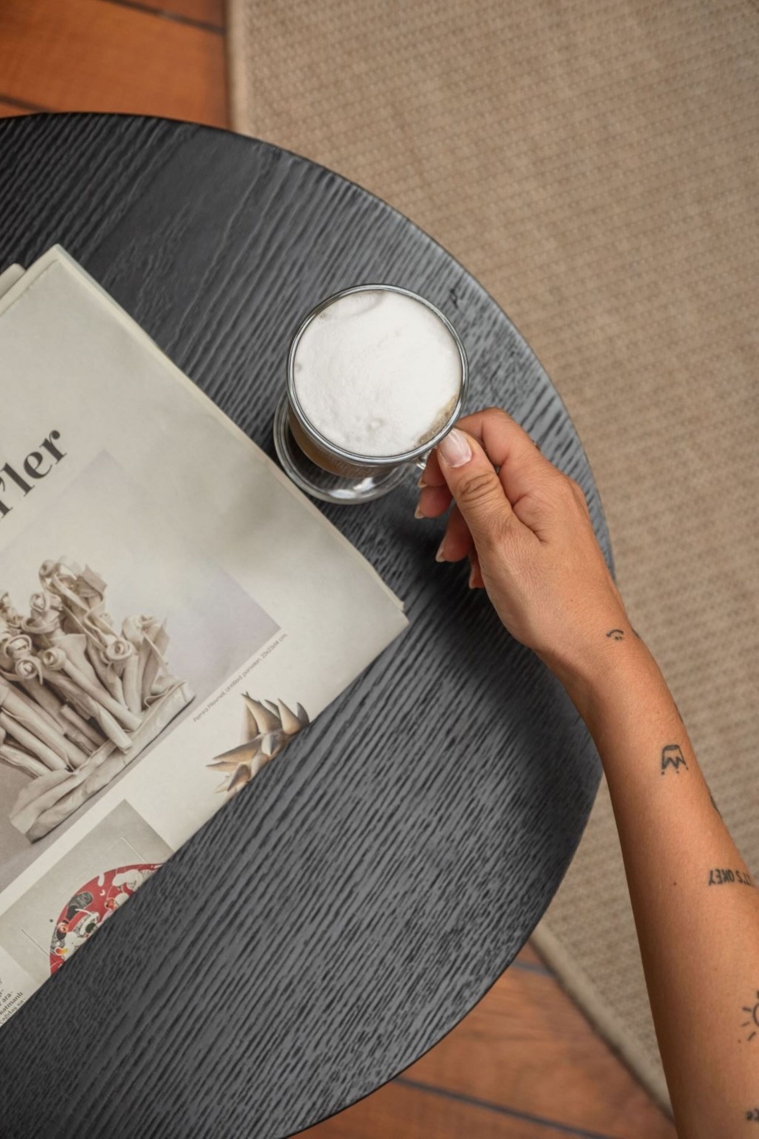 A hand holding a glass of white beverage next to a magazine on a round table.