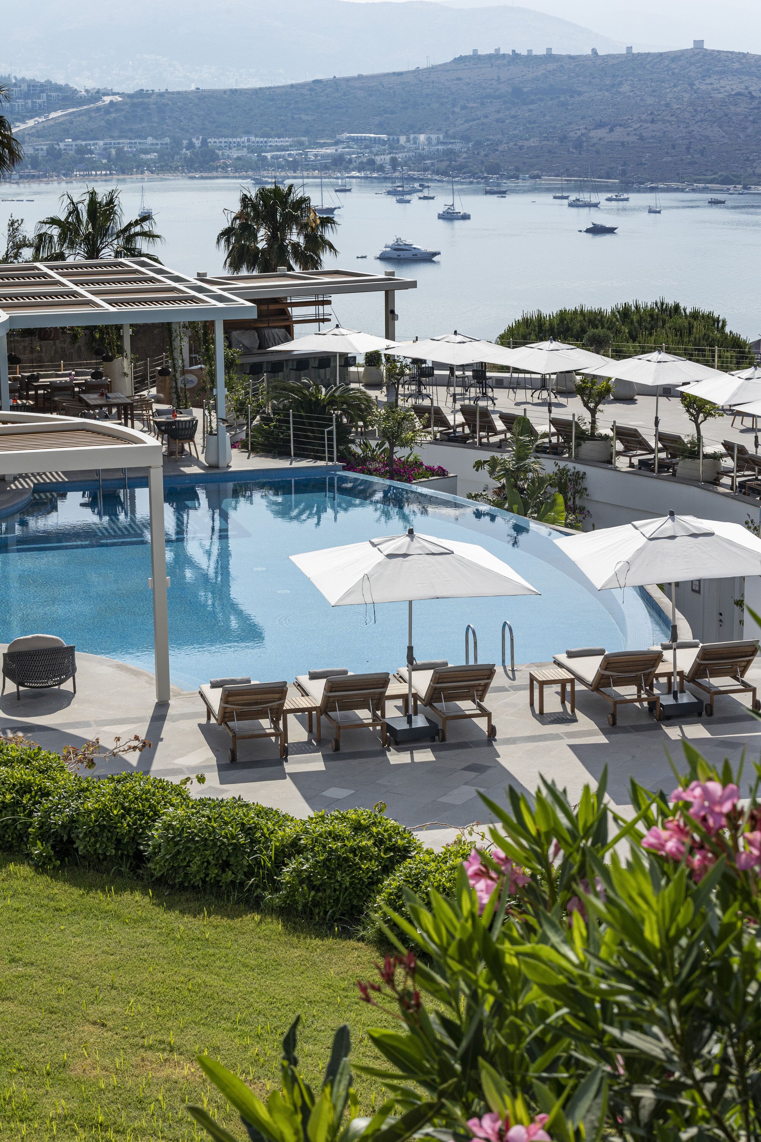 A pool area with lounge chairs and umbrellas, surrounded by greenery and overlooking a body of water with boats.