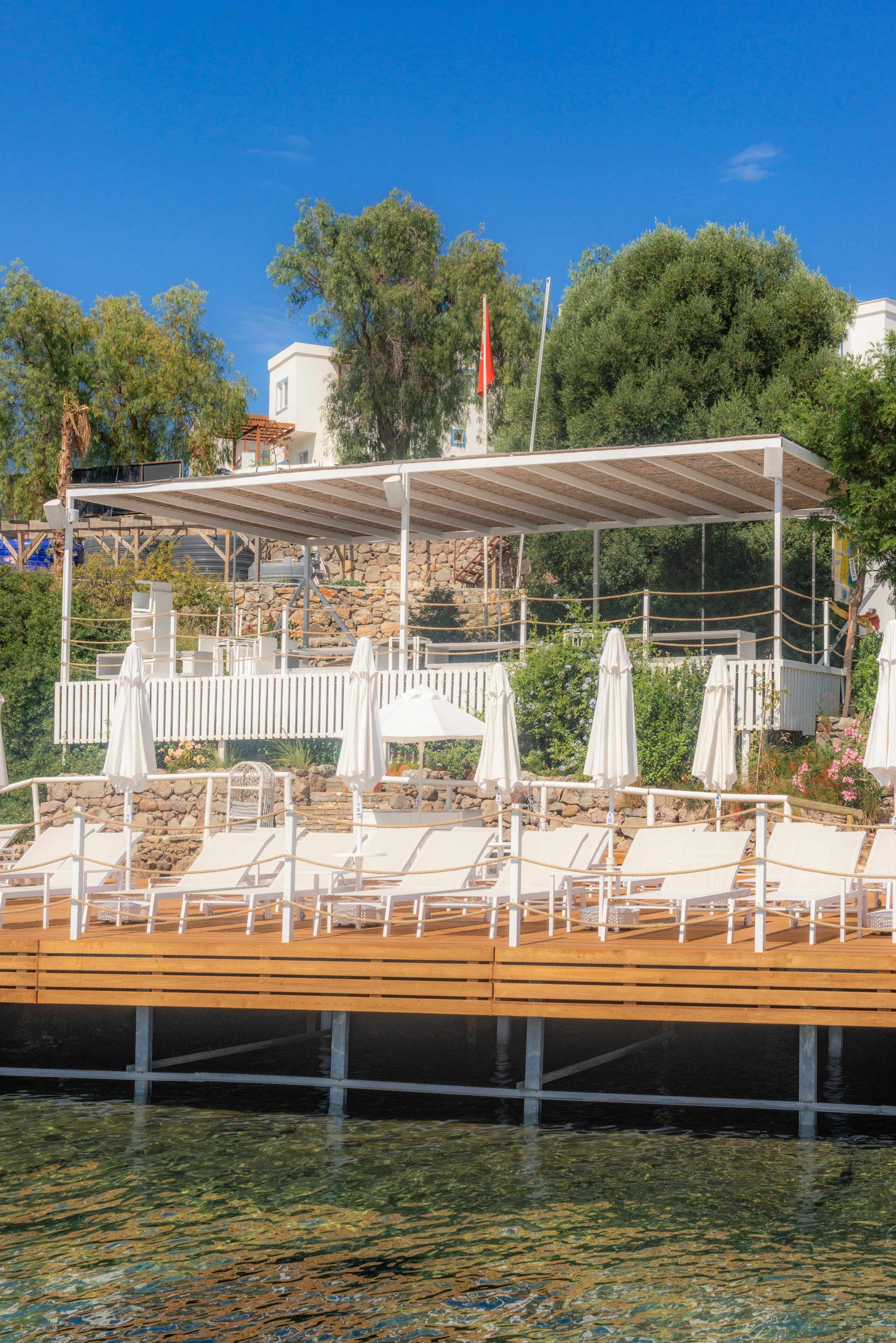 A wooden dock with lounge chairs and umbrellas, a covered seating area, and a stone wall in the background.