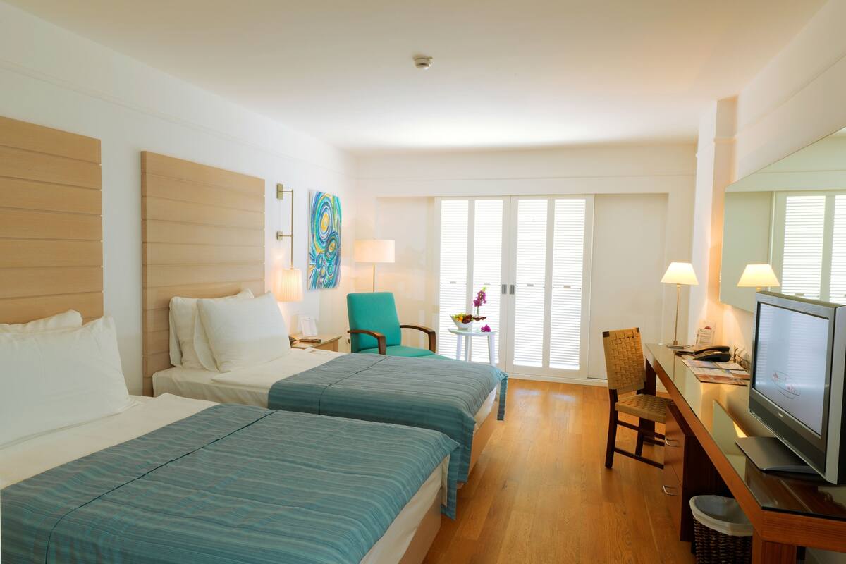 Une chambre d’hôtel avec deux lits, un bureau avec une télévision, une chaise, une petite table avec un vase de fleurs et une grande fenêtre avec des stores.
