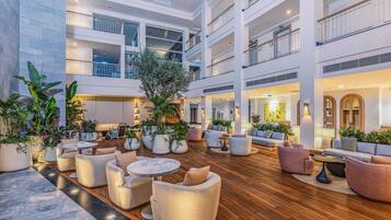 Um amplo átrio interno com vários níveis, apresentando uma área de estar central com sofás e mesas, cercada por plantas em vasos e um piso de madeira.