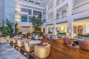 Um amplo átrio interno com vários níveis, apresentando uma área de estar central com sofás e mesas, cercada por plantas em vasos e um piso de madeira.
