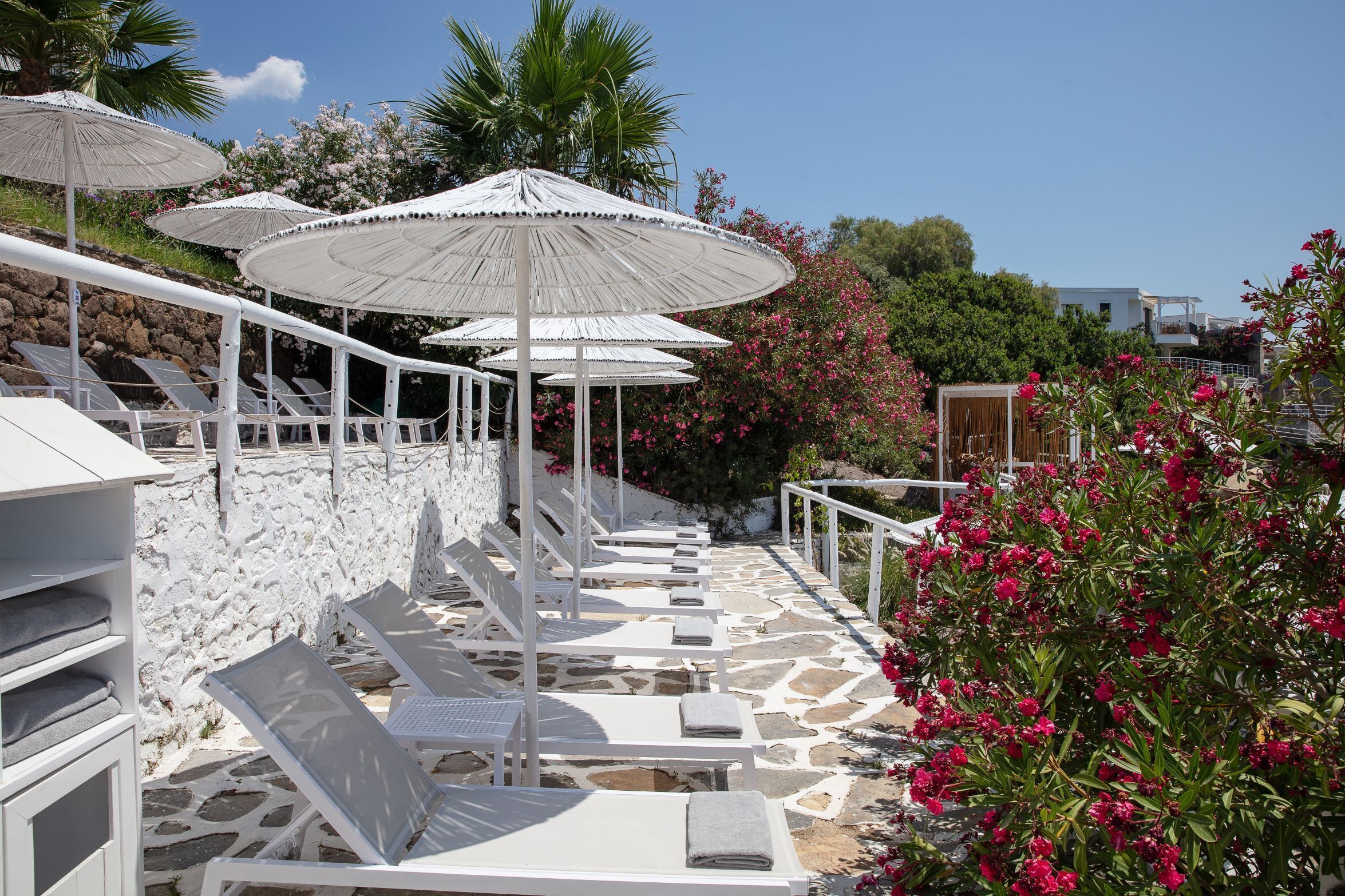 Zona relax esterna illuminata dal sole, con poltrone bianche e ombrelloni, circondata da pareti in pietra e piante fiorite.