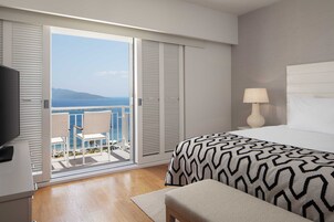 Habitación de hotel con balcón, cama con ropa de cama estampada, mesita de noche con lámpara y televisor sobre un mueble.