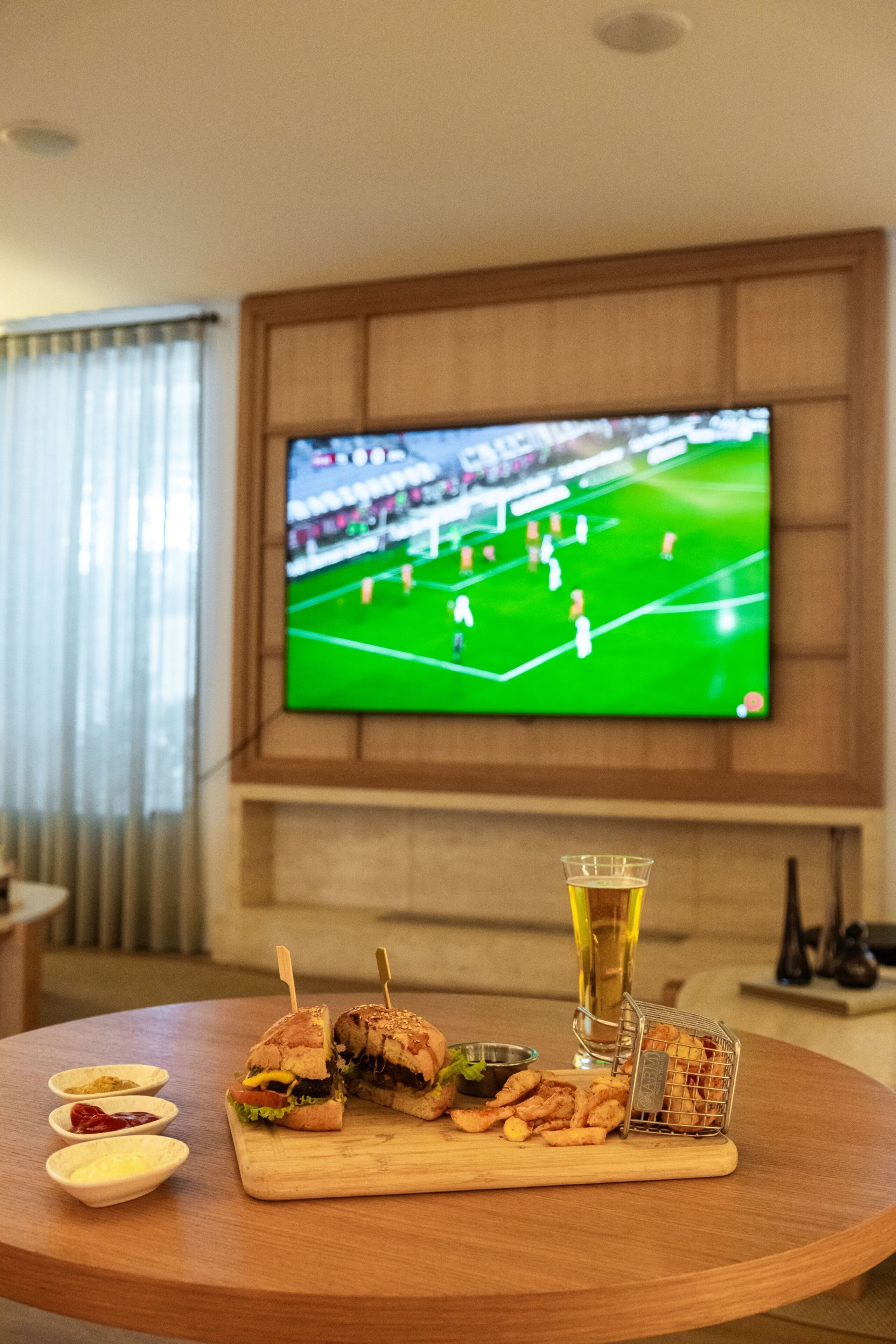 A dining table with a sandwich cut in half, a glass of beer, and small bowls of condiments. A television displaying a soccer game is mounted on the wall in the background.