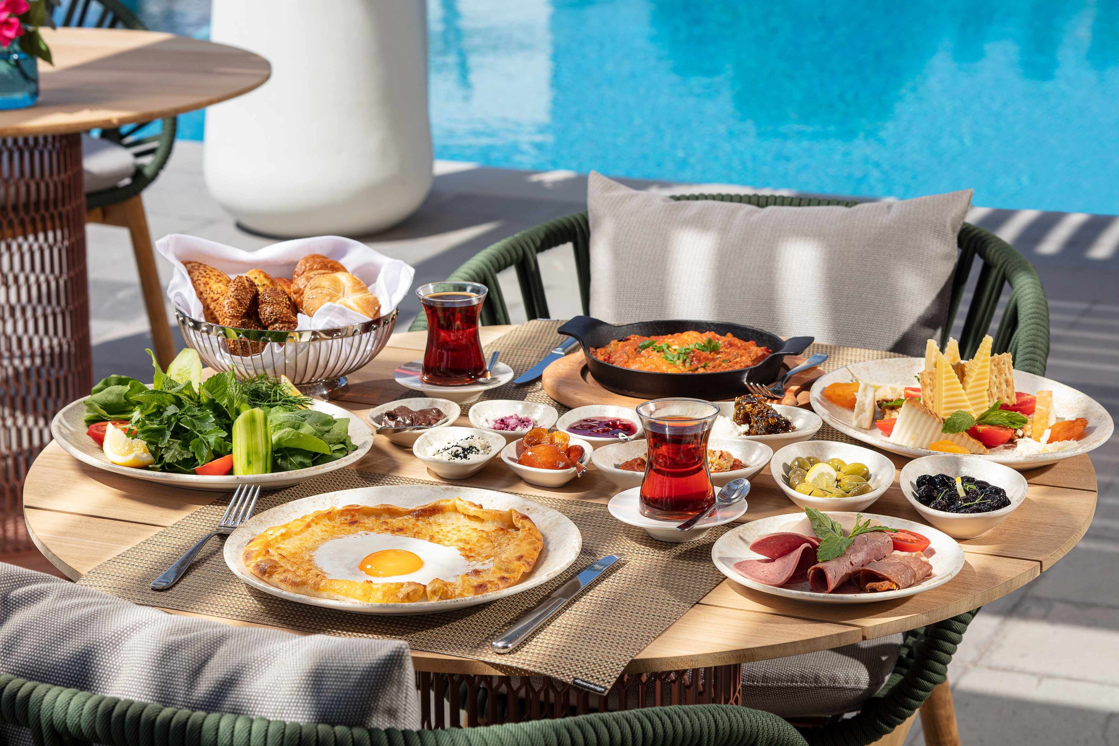 A dining table with various dishes, including a salad, a bowl of bread, and a plate of meat, set outdoors by a pool.