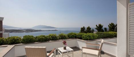A balcony with two wicker chairs, a small table with a vase, and a view of the sea and distant mountains.