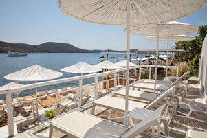 Una terraza frente al mar con sillas reclinables blancas y sombrillas, con vista a un mar tranquilo y colinas lejanas.
