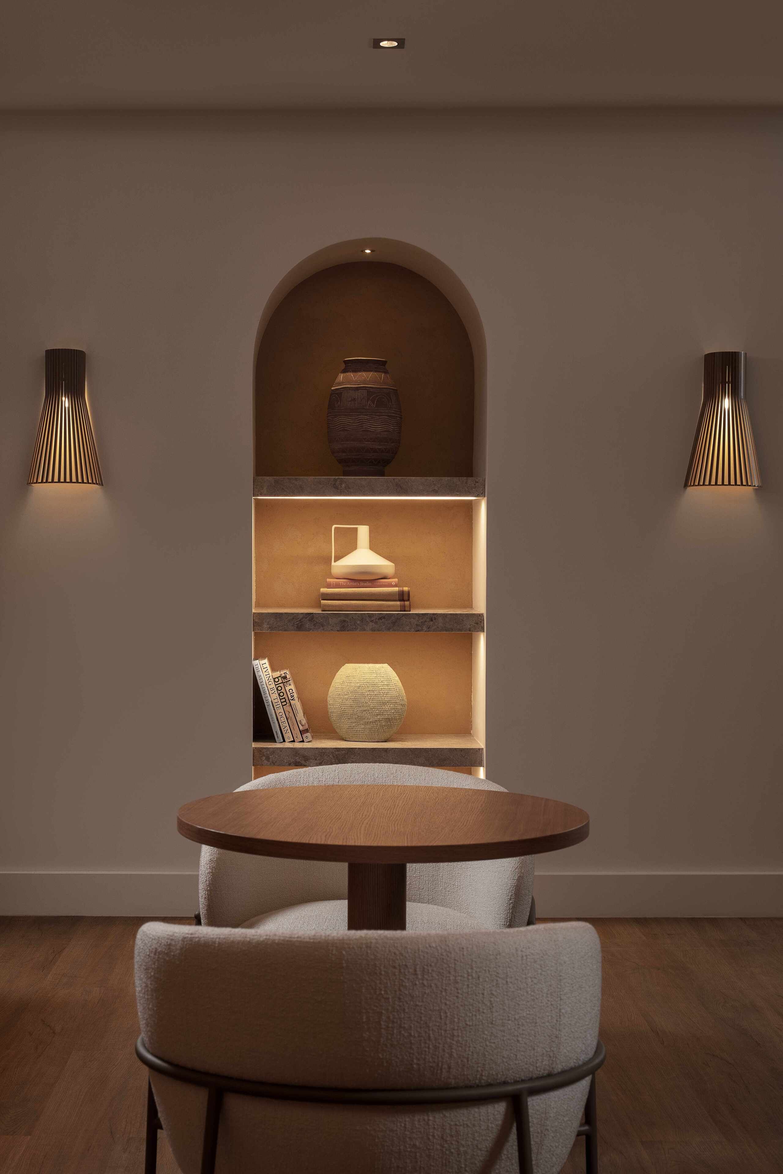 Un interior moderno con una mesa redonda y dos sillas, una estantería con objetos decorativos y dos lámparas de pared.