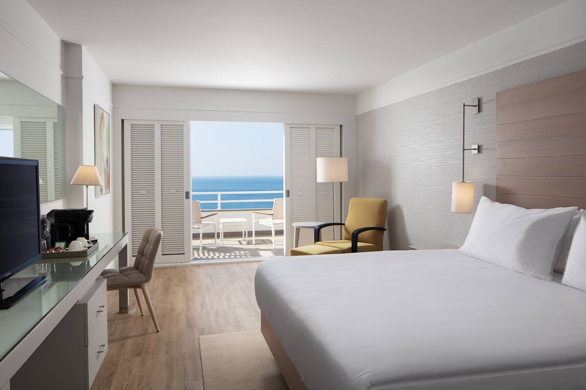 Une chambre d’hôtel avec un lit, un bureau équipé d’un ordinateur, une chaise, une lampe et une vue sur l’océan.